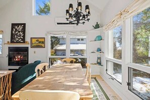 Un comedor con mesa de madera, sillas, chimenea y ventanas grandes.