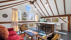 A spacious loft with a high ceiling, large windows, a kitchen area, and a cozy seating area with a red sofa and patterned rug. - Juniper Presents: The Loft at Historic 33 Acre Berrywood Farm (Three Oaks)