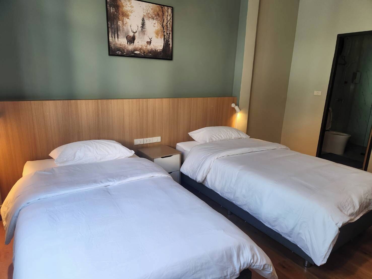 A hotel room with two single beds, a wooden headboard, a nightstand, a wall-mounted light, and a painting of deer in a forest.