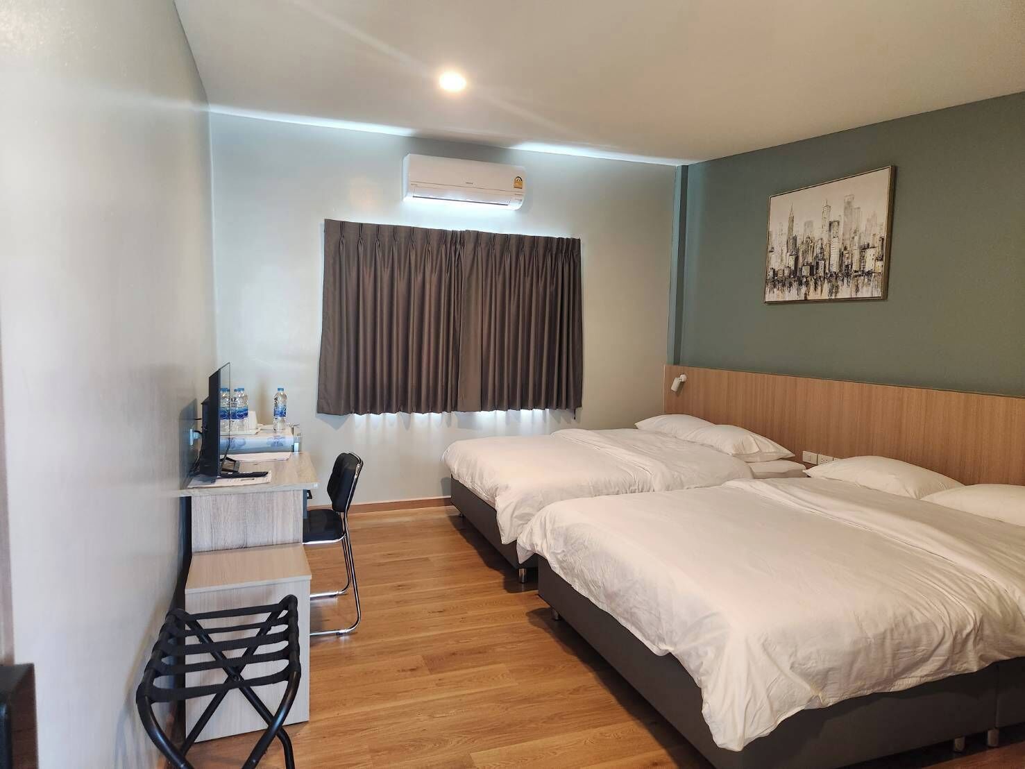 A hotel room with two beds, a desk, a chair, an air conditioner, and a cityscape painting on the wall.