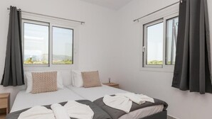 Ein Doppelbett mit grauem Kopfteil, weißer Bettwäsche und zwei Kissen. Zwei Nachttische mit Lampen, ein Fenster mit Vorhängen und Blick auf Grünfläche im Freien.