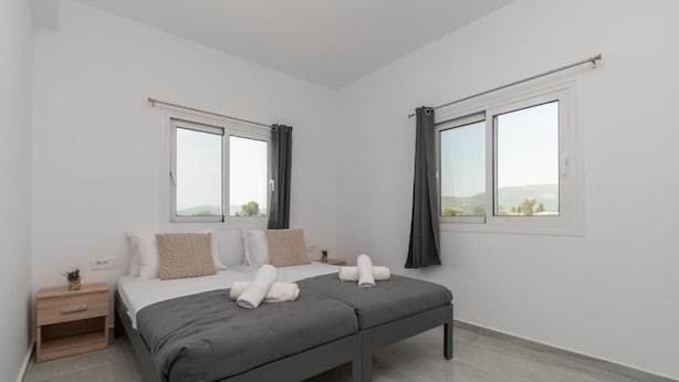 Ein modernes Schlafzimmer mit einem Bett, zwei Fenstern, einem Nachttisch und Blick ins Freie.