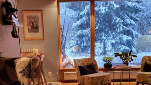 Uma sala de estar acolhedora com piso de madeira, uma janela grande com vista para a neve e uma lareira.
