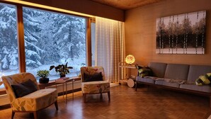 Uma sala de estar aconchegante, com uma janela grande, um sofá, duas poltronas, uma mesa com abajur e um quadro na parede.