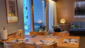 Uma área de refeições com mesa e cadeiras de madeira, luminária pendente e vista através de grandes janelas.