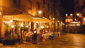 Un café al aire libre con mesas y sillas, rodeado de edificios con balcones y una torre del reloj al fondo.