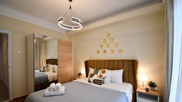 A hotel room with a large bed, a mirror, a wooden headboard, a bedside table with a lamp, and decorative leaf wall art. - Porto Sea View Apartments - Tsimiski 78 (Thessaloniki)