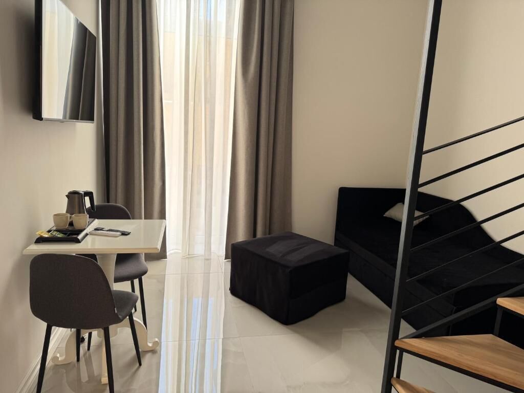 A modern hotel room with a bed, a desk with chairs, a small table with cups and a book, and a staircase.