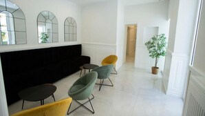 A modern living room with a black sofa, three armchairs, and a coffee table. There are three large mirrors on the wall, a potted plant, and a door leading to another room. - Gentile Suite Decumani (Naples)