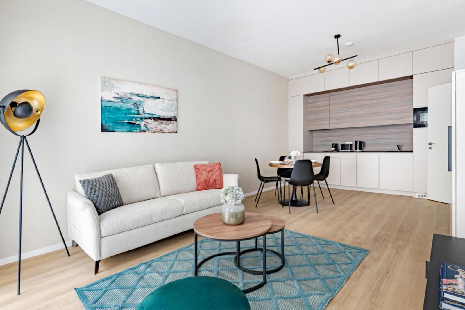 A modern living room with a sofa, coffee table, and chairs. There is a dining area with a table and chairs. The room has a kitchen with cabinets and a countertop. A floor lamp and a painting are visible.