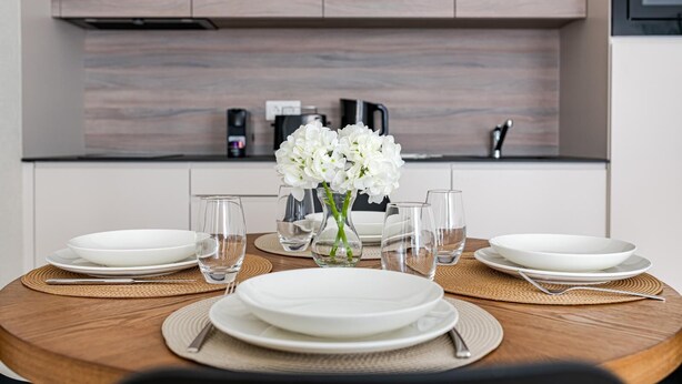 Una mesa de comedor dispuesta con platos blancos, vasos transparentes y un jarrón de flores blancas.