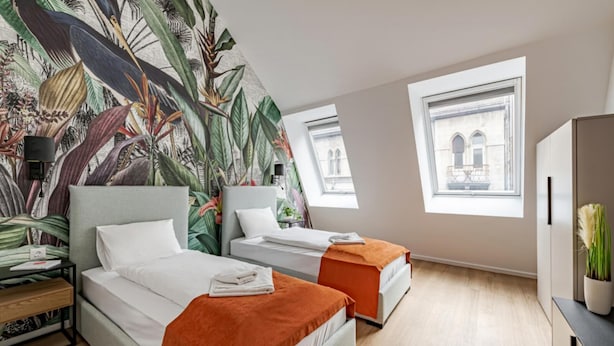 Una habitación de hotel moderna con dos camas, un gran mural tropical y ventanas amplias con vistas a la ciudad.