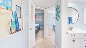 Un pasillo con un mural alusivo a la playa, una puerta blanca, una cama con colcha azul y blanca a rayas, y un baño con un espejo redondo y un lavabo.