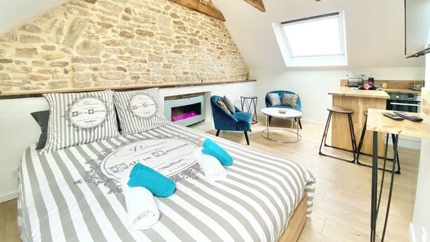 A bedroom with a stone wall, a bed with striped bedding, two chairs, a small table, and a kitchen area with a TV. - 4★ Bright Studio on Top Floor, Wifi, 5 min Walk to Quimper Station (Quimper)