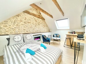Un dormitorio con pared de piedra, una cama con ropa de cama a rayas, dos sillas, una mesita y una zona de cocina con televisor.
