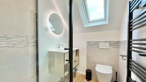 A modern bathroom with a glass shower enclosure, a round mirror with integrated lighting, a toilet, and a window above the sink. - 4★ Bright Studio on Top Floor, Wifi, 5 min Walk to Quimper Station (Quimper)
