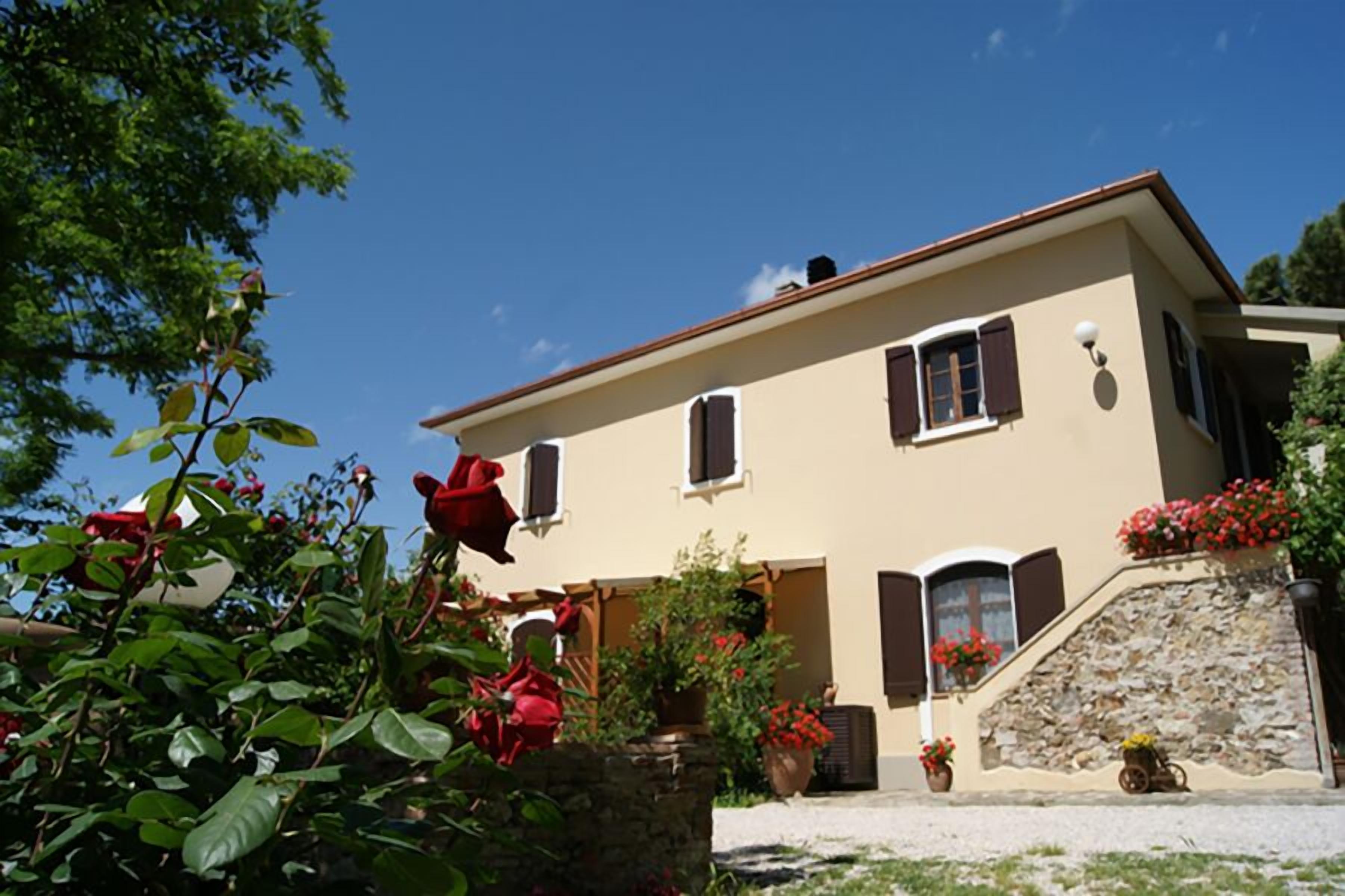 A two-story building with a stone staircase leading to the entrance, surrounded by lush greenery and red flowers.