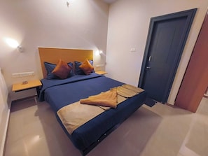 A bedroom with a bed, a nightstand, a door, and a wall-mounted light. - Apex Inn Manyata (Bengaluru)