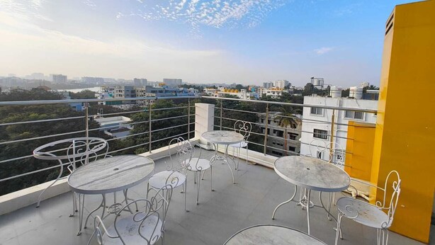 A rooftop terrace with round tables and metal chairs, overlooking a cityscape with buildings and greenery. - Apex Inn Manyata (Bengaluru)