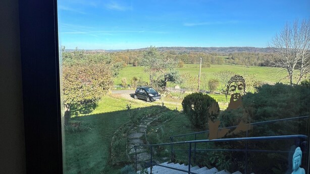 Une vue depuis une fenêtre donnant sur un paysage verdoyant, avec une statue de Bouddha et une voiture garée au loin.