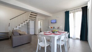 A modern living room with a white sofa, a dining table with white chairs, a staircase, and a TV mounted on the wall. - 18 Gradi Apartments (Marsala)
