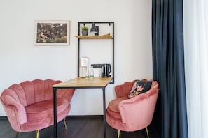 Una stanza con due sedie rosa, un tavolo in legno, uno scaffale con una pianta, un quadro incorniciato e una tenda nera.