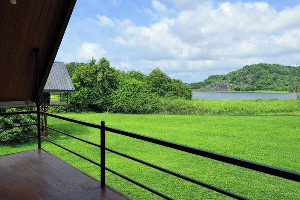 Uitzicht vanaf een balkon met houten vloer en metalen balustrade, uitkijkend over een weelderig groen veld, een waterpartij en een heuvel met bomen.
