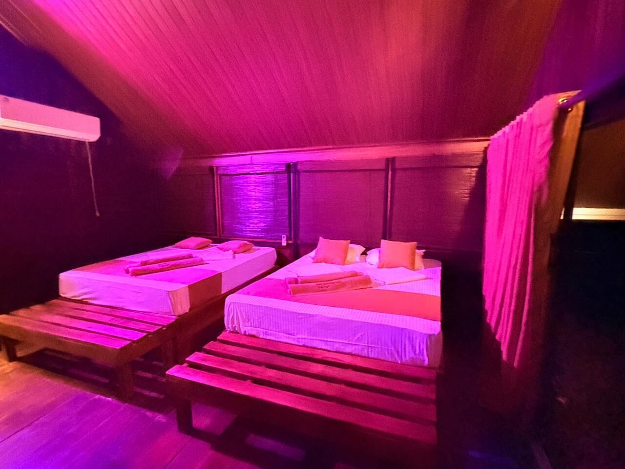 Two single beds with wooden frames and white linens, in a room with a sloped ceiling and purple lighting.