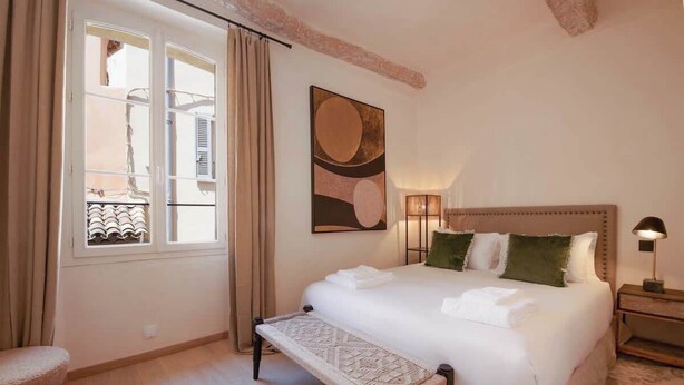 Ein Schlafzimmer mit einem großen Bett, einer Bank, einem Fenster mit Vorhängen und einem an der Wand befestigten Kunstwerk.