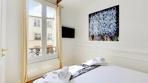 Une chambre d’hôtel avec un lit, une fenêtre donnant sur des immeubles, un tableau accroché au mur et un téléviseur à écran plat fixé au-dessus du lit.