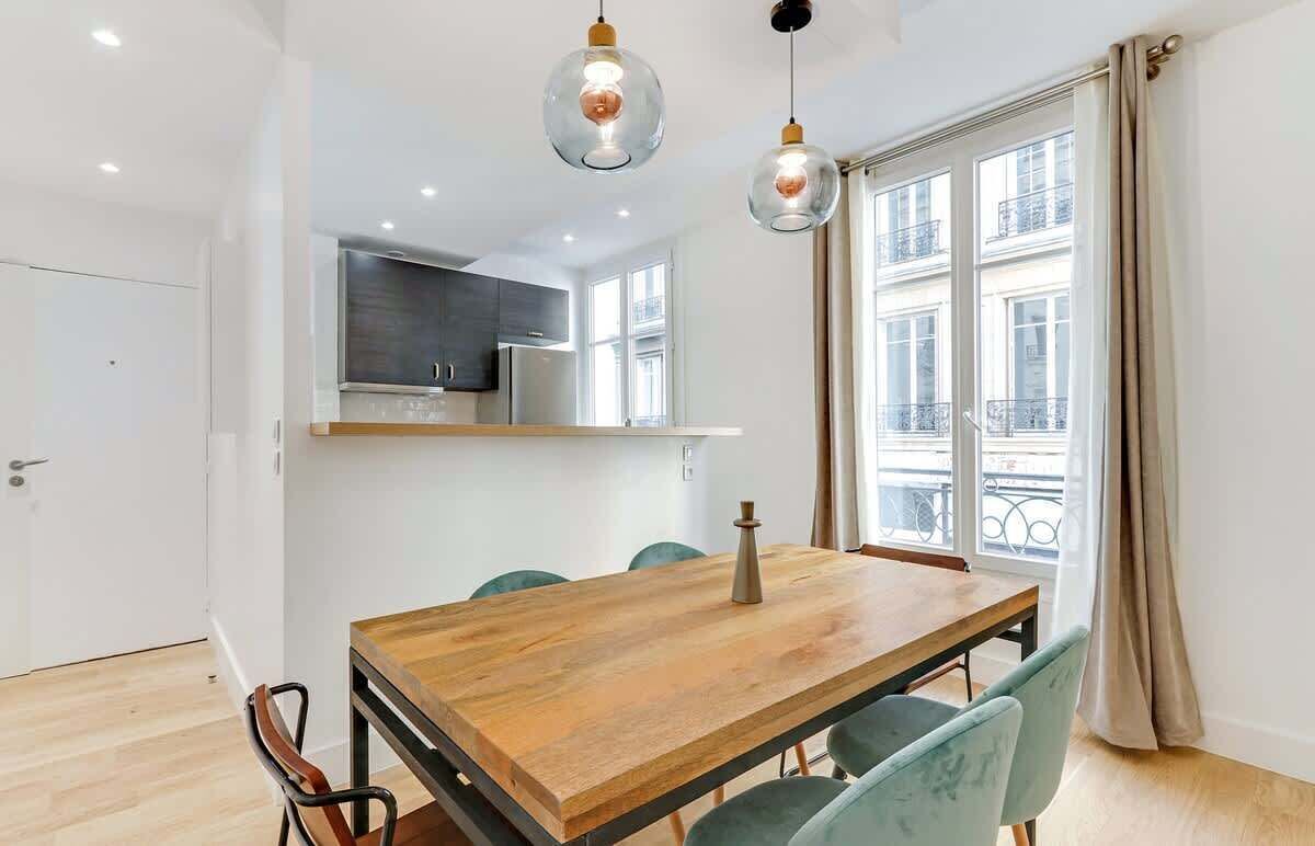 A modern dining area with a wooden table and chairs, a kitchen in the background, and large windows with curtains.