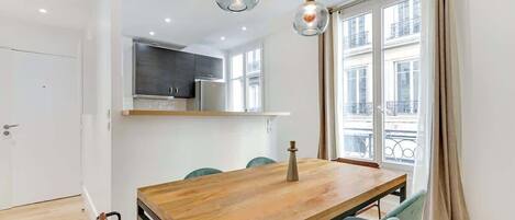 A modern dining area with a wooden table and chairs, a kitchen in the background, and large windows with curtains.