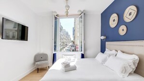 A hotel room with a bed, a chair, a TV, and a window with a city view. - An Exceptional 2-BR/2BA in Le Marais (Paris)