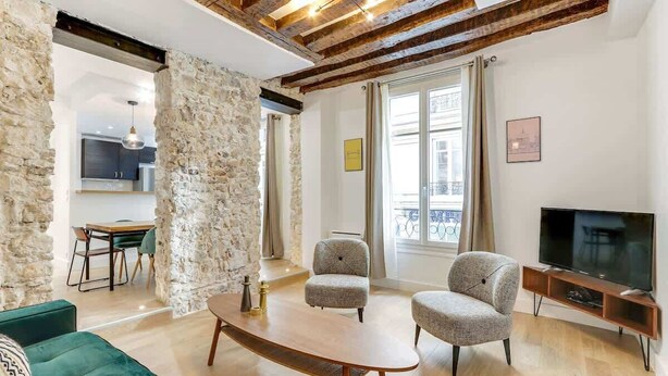 A living room with a stone wall, wooden ceiling beams, a green sofa, two armchairs, a coffee table, a TV, and a dining area in the background. - An Exceptional 2-BR/2BA in Le Marais (Paris)