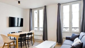 Una sala de estar moderna con un sofá, un televisor montado en la pared, una mesa de comedor con sillas y ventanas grandes con cortinas.