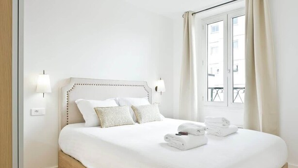 Una cama bien tendida con sábanas y almohadas blancas, una lámpara de noche y una ventana con cortinas.