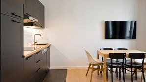 Una cocina moderna con armarios oscuros, encimera de madera y estufa empotrada. Hay una zona de comedor con una mesa y sillas, y un televisor de pantalla plana montado en la pared.