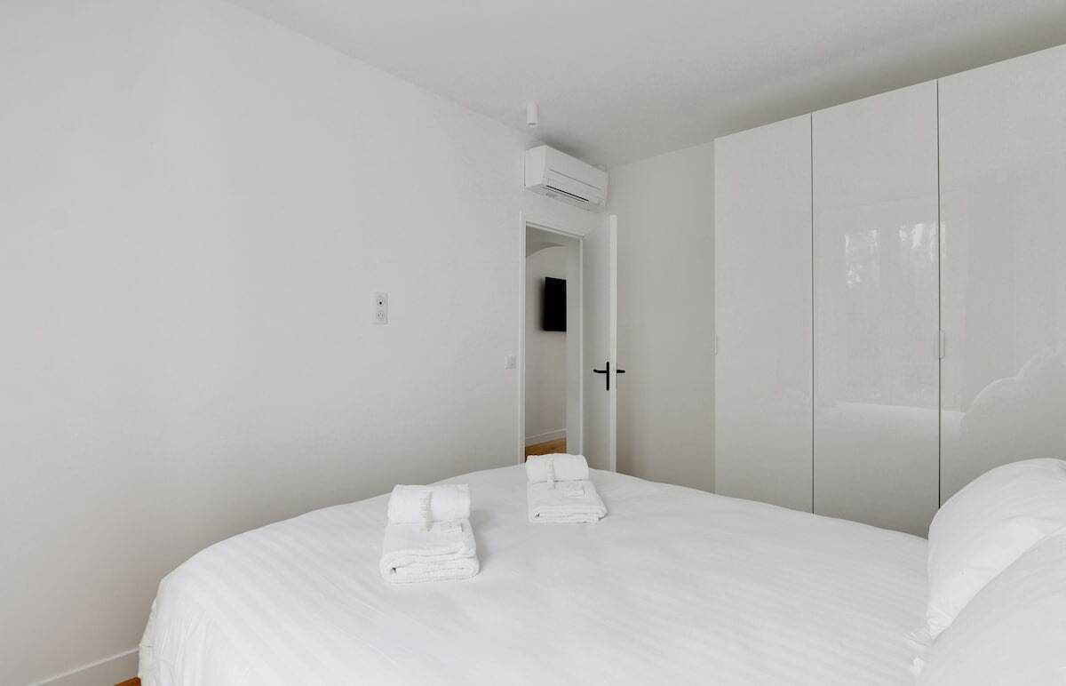 A bedroom with a bed, two folded towels, a wardrobe, and an air conditioning unit.