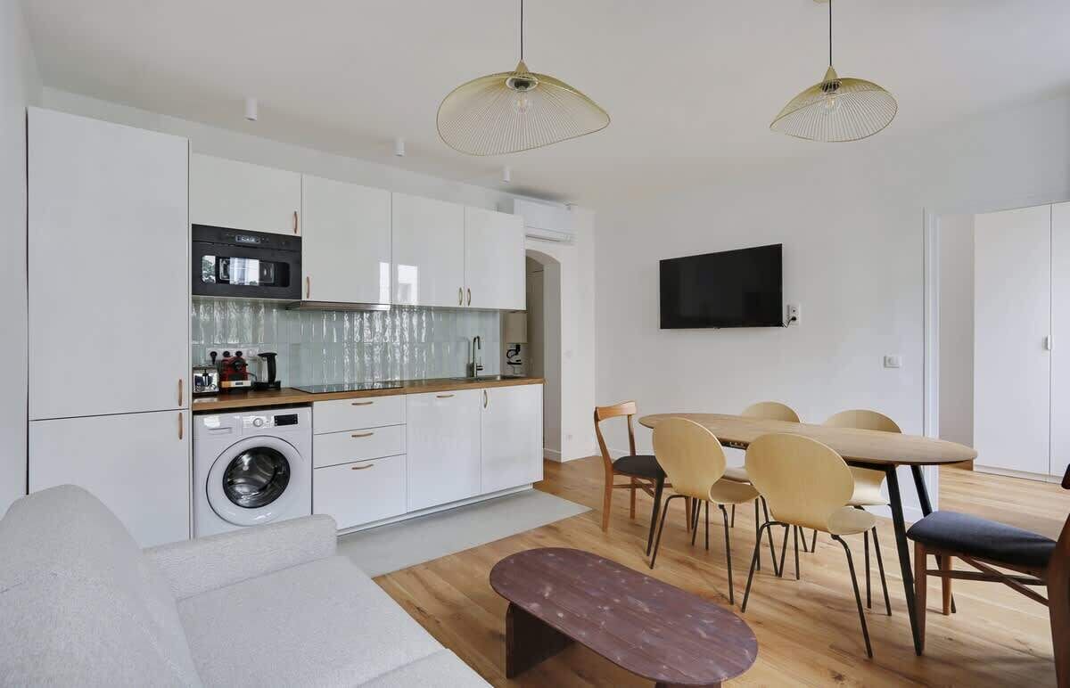 A modern kitchen with white cabinets, a built-in oven, and a washing machine. There is a dining area with a table and chairs, and a flat-screen TV mounted on the wall.