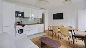 Una cucina moderna con mobili bianchi, un forno integrato e una lavatrice. C'è un'area pranzo con tavolo e sedie, e un televisore a schermo piatto montato a parete.