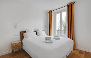 Een net opgemaakt bed met witte lakens en twee gevouwen handdoeken, een houten nachtkastje met lades en een raam met oranje gordijnen.