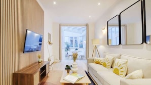 A modern living room with a flat-screen TV mounted on a wooden panel wall, a white sofa with sun-shaped pillows, a coffee table with a book and a vase, and a large mirror on the wall. - An Amazing 2-BR in Le Marais (Paris)
