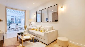 A modern living room with a sofa, coffee table, and ottoman. There are three wall-mounted mirrors and a lamp. A dining area is visible in the background. - An Amazing 2-BR in Le Marais (Paris)