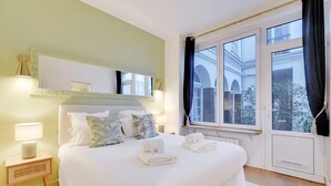 A hotel room with a large bed, a bedside table with a lamp, a mirror, and a window with curtains. - An Amazing 2-BR in Le Marais (Paris)