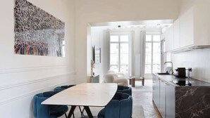 Moderni keittiö, jossa on marmoritaso, ruokailutila valkoisella pöydällä ja sinisillä tuoleilla sekä suuri abstrakti maalaus seinällä.