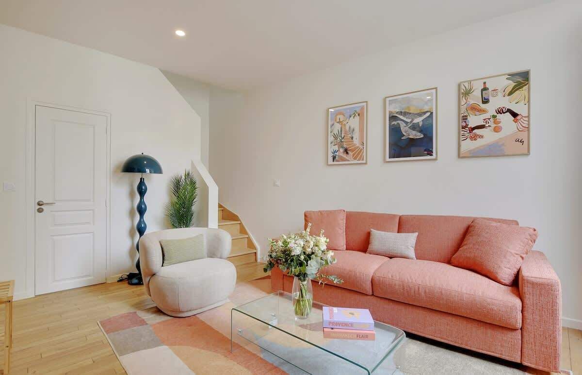 Ein Wohnzimmer mit einem rosafarbenen Sofa, einem weißen Sessel, einem Glastisch mit einer Blumenvase und einem Bücherregal. An der Wand hängen drei gerahmte Kunstwerke.