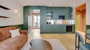 Una sala moderna con sofá y una mesa de centro, una cocina con armarios azul verdosos y un horno empotrado, y un pasillo que lleva a una habitación luminosa con radiador.