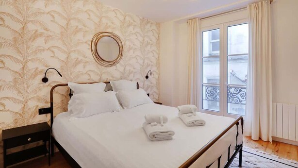 A hotel room with a bed, a round mirror, a nightstand, and a window with a view of a balcony. - A Cosy 1-BR/1BA Duplex in Montmartre (Paris)