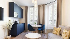 A modern living room with a sofa, a round dining table with chairs, a kitchen area with cabinets, and large windows with curtains. - A Romantic 1-BR with Services in Batignolles (Paris)