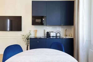 A modern kitchen with dark cabinets, a microwave, a kettle, and a toaster. There is a dining table with chairs and a vase with flowers on the counter. - A Romantic 1-BR with Services in Batignolles (Paris)